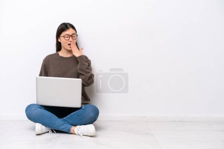 Téléchargez les photos : Jeune femme avec un ordinateur portable assis sur le sol bâillant et couvrant la bouche grande ouverte avec la main - en image libre de droit