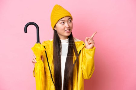 Junge Chinesin mit regenfestem Mantel und Regenschirm auf rosa Hintergrund, der auf eine großartige Idee hinweist