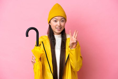 Junge Chinesin mit regendichtem Mantel und Regenschirm auf rosa Hintergrund glücklich und zählt mit den Fingern drei
