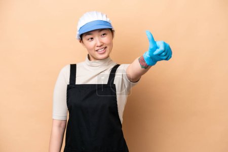 Photo for Young Chinese Fishmonger woman wearing an apron and holding a raw fish isolated on pink background giving a thumbs up gesture - Royalty Free Image