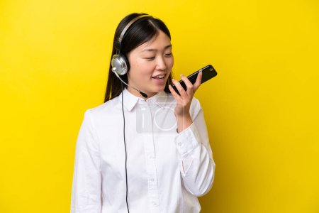 Photo for Telemarketer Chinese woman working with a headset isolated on yellow background keeping a conversation with the mobile phone - Royalty Free Image