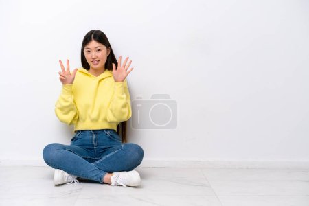 Foto de Joven mujer china sentada en el suelo aislada en la pared blanca contando ocho con los dedos - Imagen libre de derechos
