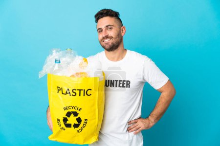 Foto de Hombre caucásico sosteniendo una bolsa llena de botellas de plástico para reciclar aislado sobre fondo azul posando con los brazos en la cadera y sonriendo - Imagen libre de derechos
