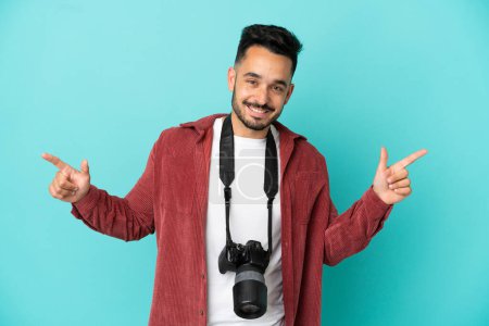 Foto de Joven fotógrafo caucásico hombre aislado sobre fondo azul señalando el dedo a los laterales y feliz - Imagen libre de derechos