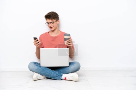 Téléchargez les photos : Adolescent russe homme tenant PC assis sur le sol isolé sur fond blanc tenant café à emporter et un mobile - en image libre de droit