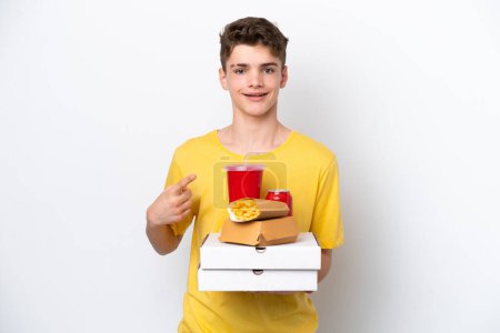 Téléchargez les photos : Adolescent homme russe tenant fast food isolé sur fond blanc pointant vers le côté pour présenter un produit - en image libre de droit