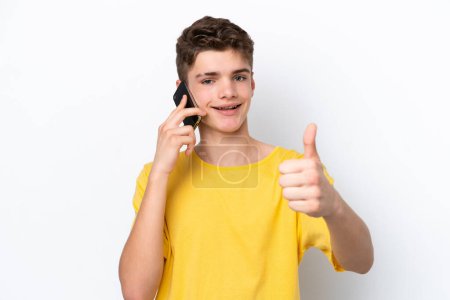 Téléchargez les photos : Homme adolescent utilisant un téléphone mobile isolé sur fond blanc avec les pouces levés parce que quelque chose de bon s'est passé - en image libre de droit