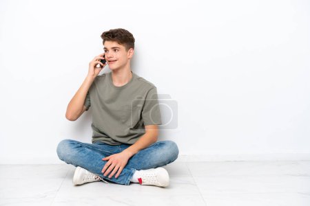 Téléchargez les photos : Adolescent homme russe assis sur le sol isolé sur fond blanc en gardant une conversation avec le téléphone mobile - en image libre de droit