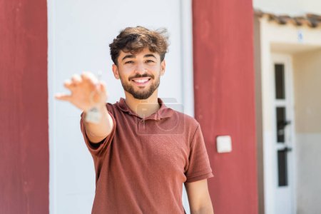Téléchargez les photos : Jeune homme beau arabe tenant les clés de la maison à l'extérieur avec une expression heureuse - en image libre de droit