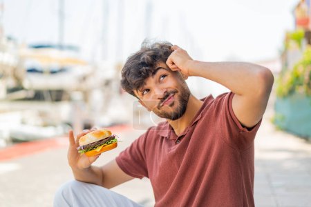 Téléchargez les photos : Jeune homme beau arabe tenant un hamburger à l'extérieur ayant des doutes et avec une expression du visage confuse - en image libre de droit