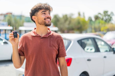 Foto de Joven árabe guapo hombre sosteniendo la llave del coche al aire libre mirando hacia arriba mientras sonríe - Imagen libre de derechos