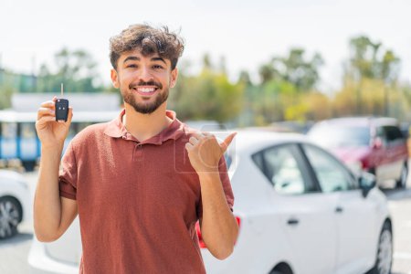 Téléchargez les photos : Jeune homme beau arabe tenant la clé de voiture à l'extérieur pointant vers le côté pour présenter un produit - en image libre de droit