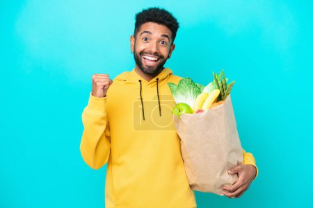 Photo for Young Brazilian man taking a bag of takeaway food isolated on blue background celebrating a victory in winner position - Royalty Free Image
