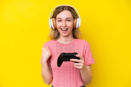 Foto de Chica joven inglesa rubia jugando con un controlador de videojuegos aislado sobre fondo amarillo celebrando una victoria en posición ganadora - Imagen libre de derechos
