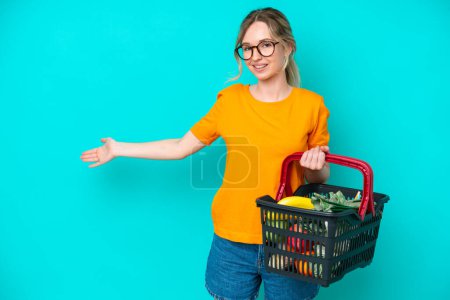 Foto de Blonde English young girl holding a shopping basket full of food isolated on blue background extending hands to the side for inviting to come - Imagen libre de derechos