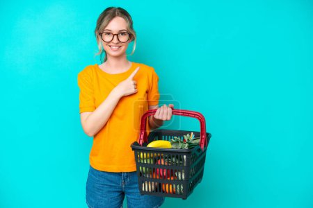 Photo for Blonde English young girl holding a shopping basket full of food isolated on blue background pointing to the side to present a product - Royalty Free Image