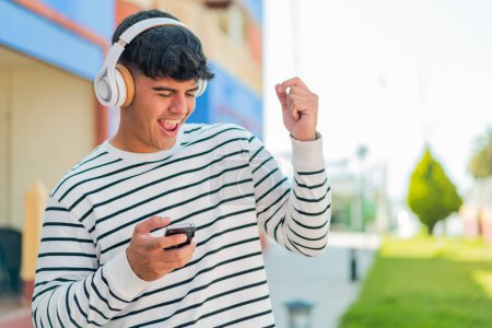 Téléchargez les photos : Jeune homme hispanique à l'extérieur écouter de la musique et faire un geste de guitare - en image libre de droit