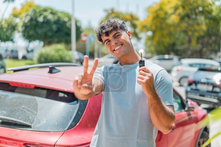 Téléchargez les photos : Jeune hispanique avec des clés de voiture souriant et montrant signe de victoire - en image libre de droit