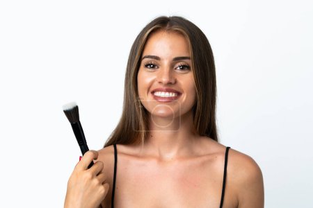 Photo for Young Uruguayan woman isolated on white background holding makeup brush and whit happy expression - Royalty Free Image