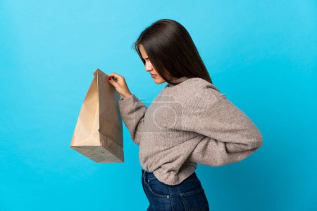 Téléchargez les photos : Femme prenant un sac de nourriture à emporter isolé sur fond bleu souffrant de maux de dos pour avoir fait un effort - en image libre de droit