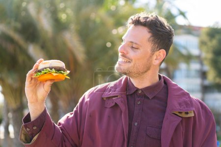 Photo for Young caucasian man holding a burger at outdoors with happy expression - Royalty Free Image