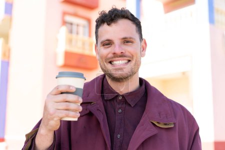 Téléchargez les photos : Jeune homme caucasien à l'extérieur tenant un café à emporter avec expression heureuse - en image libre de droit