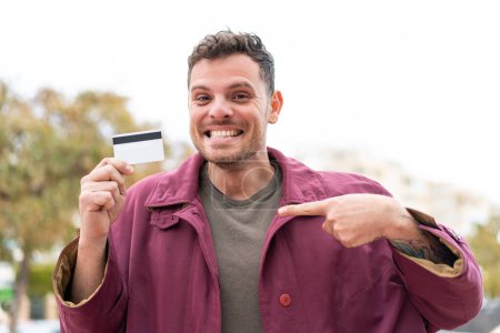Téléchargez les photos : Jeune homme caucasien tenant une carte de crédit à l'extérieur avec expression faciale surprise - en image libre de droit