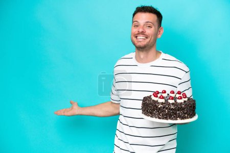 Foto de Joven hombre caucásico sosteniendo pastel de cumpleaños aislado sobre fondo azul extendiendo las manos a un lado para invitar a venir - Imagen libre de derechos