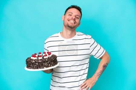 Foto de Joven hombre caucásico sosteniendo pastel de cumpleaños aislado sobre fondo azul posando con los brazos en la cadera y sonriendo - Imagen libre de derechos
