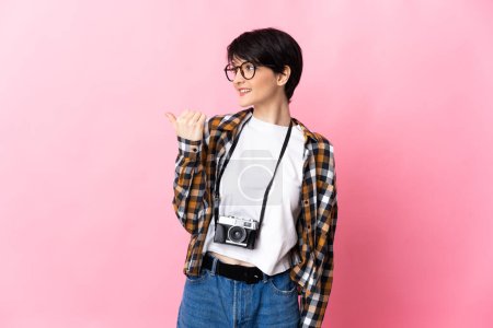 Téléchargez les photos : Jeune photographe femme isolée sur fond rose pointant vers le côté pour présenter un produit - en image libre de droit