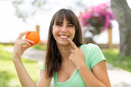 Téléchargez les photos : Jeune femme à l'extérieur tenant une orange avec une expression heureuse - en image libre de droit