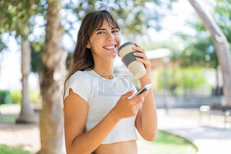 Téléchargez les photos : Jeune femme à l'extérieur en utilisant un téléphone portable et tenant un café avec une expression heureuse - en image libre de droit