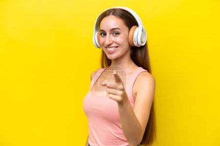 Téléchargez les photos : Jeune femme caucasienne isolée sur fond jaune écoutant de la musique et pointant vers l'avant - en image libre de droit