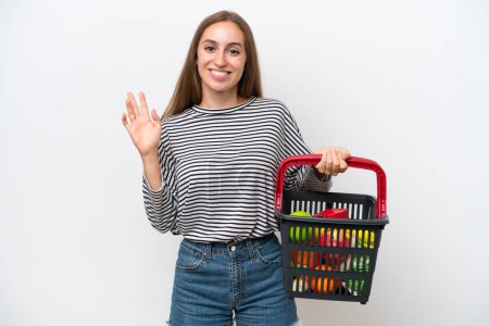 Photo for Young Rumanian woman holding a shopping basket full of food isolated on white background saluting with hand with happy expression - Royalty Free Image