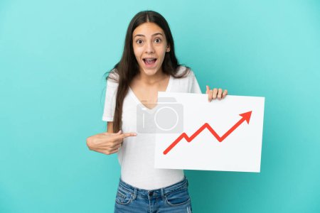 Photo for Young French woman isolated on blue background holding a sign with a growing statistics arrow symbol with surprised expression - Royalty Free Image