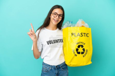 Téléchargez les photos : Jeune fille française tenant un sac rempli de bouteilles en plastique pour recycler montrant et levant un doigt en signe de la meilleure - en image libre de droit