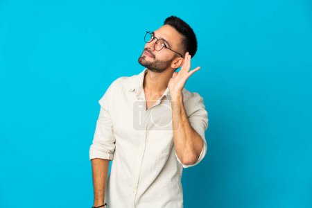 Photo for Young caucasian man isolated on blue background listening to something by putting hand on the ear - Royalty Free Image