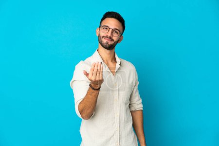 Photo for Young caucasian man isolated on blue background inviting to come with hand. Happy that you came - Royalty Free Image