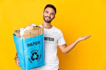 Foto de Joven hombre caucásico sosteniendo una bolsa de reciclaje llena de papel para reciclar aislado sobre fondo blanco extendiendo las manos a un lado para invitar a venir - Imagen libre de derechos
