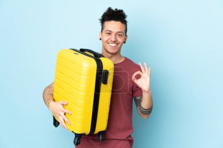 Photo for Caucasian man over isolated blue background in vacation with travel suitcase and making OK sign - Royalty Free Image