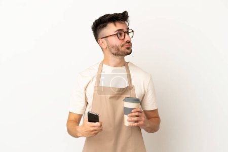 Téléchargez les photos : Serveur restaurant homme caucasien isolé sur fond blanc tenant café à emporter et un mobile tout en pensant quelque chose - en image libre de droit