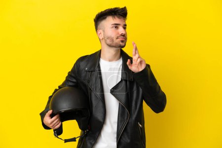 Foto de Joven hombre caucásico con casco de moto aislado sobre fondo amarillo con dedos cruzando y deseando lo mejor - Imagen libre de derechos
