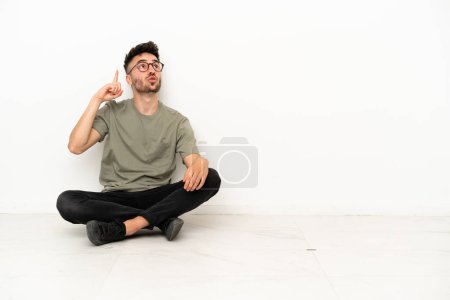 Téléchargez les photos : Jeune homme caucasien assis sur le sol isolé sur fond blanc pensant une idée pointant le doigt vers le haut - en image libre de droit