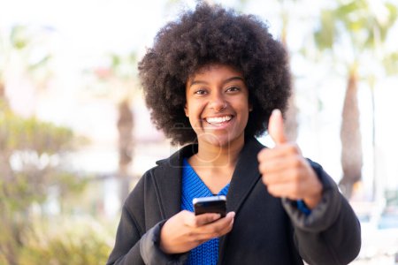 Foto de Chica afroamericana al aire libre usando el teléfono móvil mientras hace pulgares hacia arriba - Imagen libre de derechos