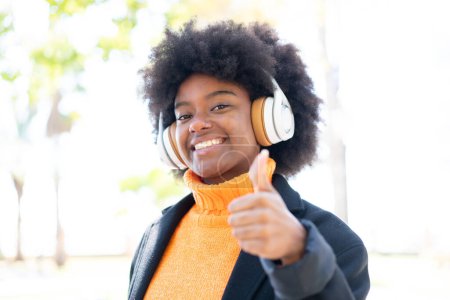Téléchargez les photos : Afro-américaine fille à l'extérieur écouter de la musique et avec pouce vers le haut - en image libre de droit