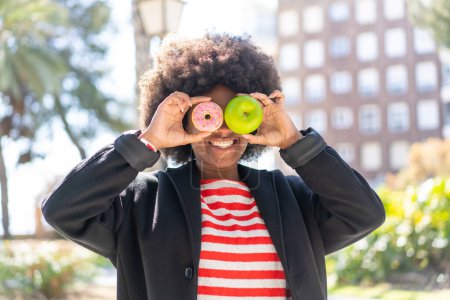 Téléchargez les photos : Afro-américaine fille à l'extérieur tenant pomme et beignet avec expression heureuse - en image libre de droit