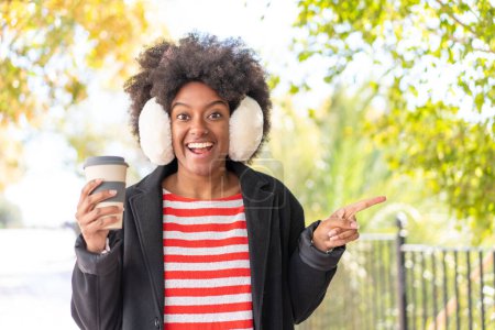 Photo for African American girl wearing winter muffs while holding a coffee at outdoors surprised and pointing finger to the side - Royalty Free Image