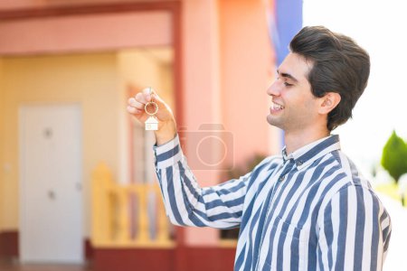 Foto de Joven hombre guapo sosteniendo las llaves de casa al aire libre con expresión feliz - Imagen libre de derechos