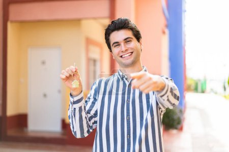Téléchargez les photos : Jeune homme beau tenant les clés de la maison à l'extérieur pointe du doigt vers vous avec une expression confiante - en image libre de droit