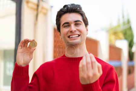 Foto de Joven hombre guapo sosteniendo un Bitcoin al aire libre invitando a venir con la mano. Feliz de que hayas venido. - Imagen libre de derechos